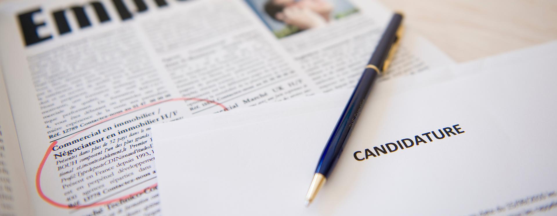 extrait d'un journal avec une offre d'emploi entourée de rouge, un stylo et une feuille sur laquelle est noté le mot candidature